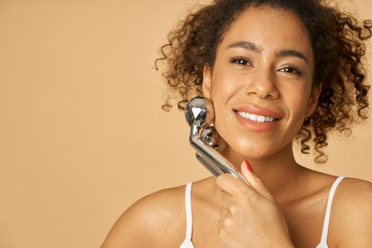 Close up portrait of beautiful young woman smiling at camera while using silver metal face roller, posing isolated over beige background. Facial massage concept