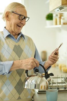 Excited elderly gentleman smiling in the middle a video-call and making himself a cup of tea