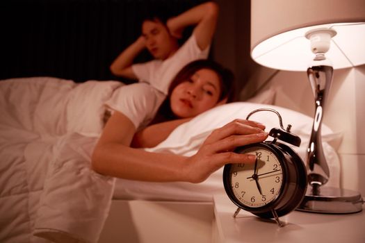 sleepy young couple in bed with extending hand to alarm clock in the morning