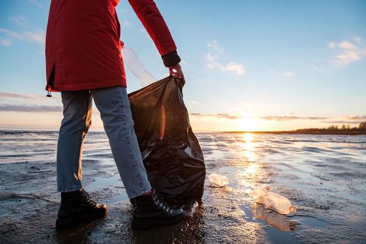 A woman cleans the bank of plastic bottles and puts the trash in a garbage bag. Environmental pollution by plastic. Recyclable packaging.