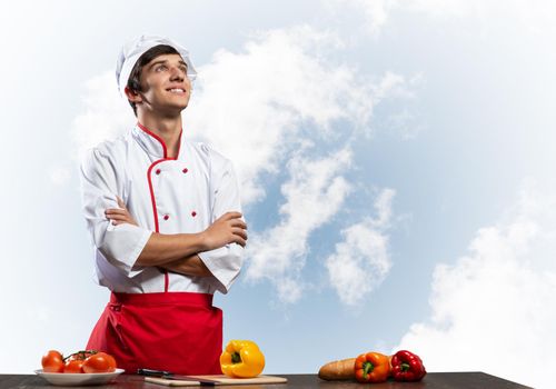 Young male chef standing with folded arms near cooking table with vegetables. Handsome chef in white hat and red apron on blue sky background. Cooking classes advertising. Vegetarian food concept