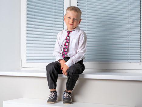 A cute little boy sits near a window closed by horizontal white blinds on the windowsill. It is possible to use the concept for advertising plastic windows and blinds.