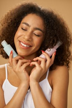Lovely young mixed race woman looking pleased, standing with eyes closed and holding two bottles with beauty product isolated over beige background. Skincare concept