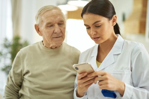 Waist up of elderly man and female doctor in white coat standing and using phone while reading message