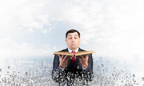 Surprised businessman looking into open old book. Startled man in business suit and tie standing on downtown background with flying various letters. Business education and knowledges concept.