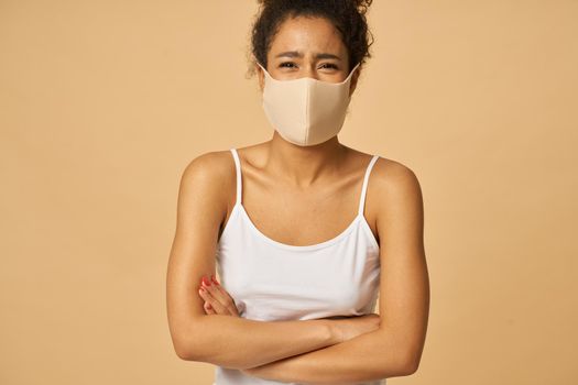 Joyful young mixed race woman wearing protective facial mask looking at camera while posing with arms crossed isolated over beige background. Safety, pandemic concept