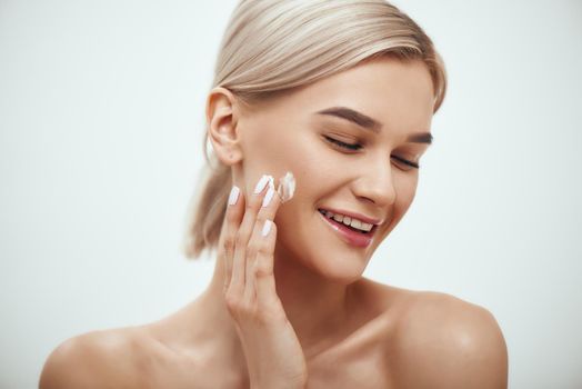 Portrait of cute blonde woman spreading cream on her face and smiling while standing against grey background. Studio shot. Skin care. Beauty concept. Cosmetics concept