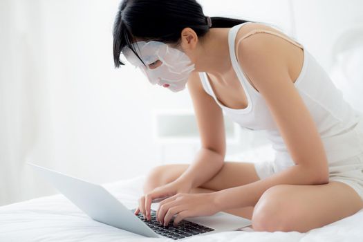 Beautiful young asian woman with sheet facial mask sitting working on laptop computer on bed at bedroom, beauty girl applying face mask for skincare of wrinkle at home, skin care, health and wellness.