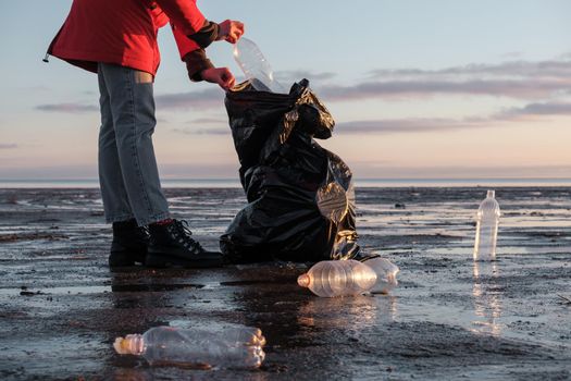 A woman cleans the bank of plastic bottles and puts the trash in a garbage bag. Environmental pollution by plastic. Recyclable packaging.