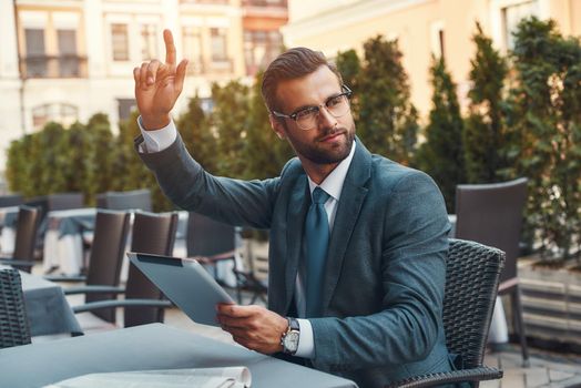 Portrait of handsome bearded businessman in eyeglasses holding touchpad and calling waiter while sitting in restaurant outdoors. Digital concept. Business concept