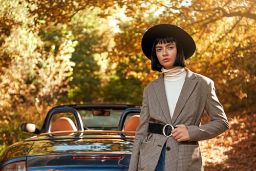 Attractive young woman posing near cabriolet. She wears black hat and sunglasses. Stylish autumn.