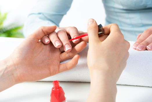Manicurist working with client's nails at table. Closeup of beautiful female hands applying red nail polish in beauty salon. Professional nail care and beautician procedure in beauty center.