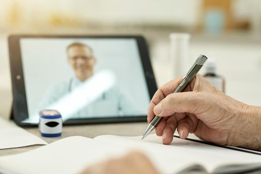 Selective focius photo of person with pen writing down notes while contacting a doctor via a video call