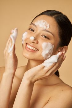Beauty portrait of joyful young woman smiling while applying gentle foam facial cleanser isolated over beige background. Beauty products and skin care concept