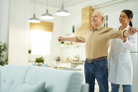 Happy female doctor in white coat helping an elderly man to do exercises in his apartment