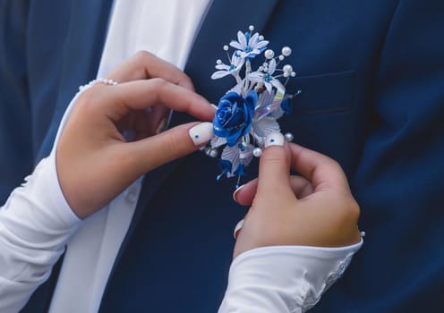 Hands of the bride in wedding gloves touch the decoration in the form of a rose flower on a blue suit of the groom, close-up.