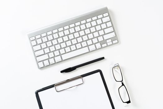 Top view of well organized workplace with office accessories. Clipboard with blank paper page, glasses and wireless keyboard on white surface. Education, creativity and working concept with copy space