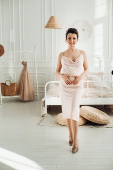 Full-length photo of a pregnant brunette lady in beige dress touching her belly and looking at the camera