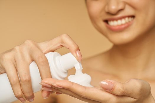 Cropped shot of young woman smiling, holding a bottle of gentle foam facial cleanser isolated over beige background. Beauty products and skin care concept