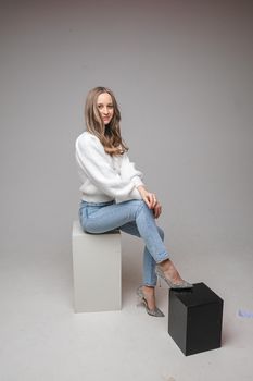 Studio full length portrait of pretty blonde woman in light blue jeans, fashionable heels and white pullover sitting on white cube and leaning on black with her leg.