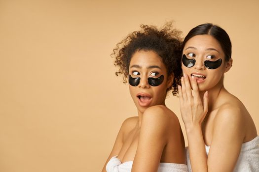 Two playful young women wrapped in towels making suprised facial expression, posing with applied black hydro gel under eye patches isolated over beige background. Skincare routine concept