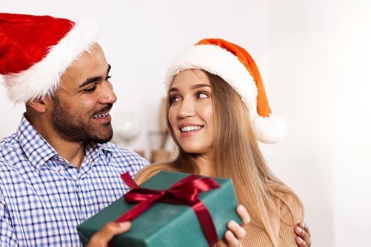 Romantic multiethnic couple in Christmas hats exchanging gifts, close up portrait