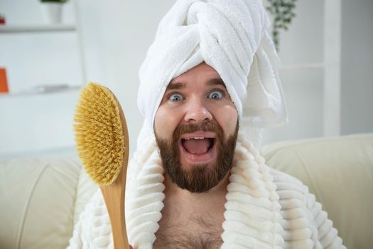 Portrait of cheerful guy wear turban towel holding in hands massage brush while sits on sofa. Male skin care and spa concept