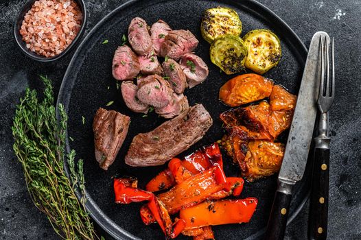 Roasted lamb or goat tenderloin Meat steak in plate with grilled vegetables. Black wooden background. Top view.