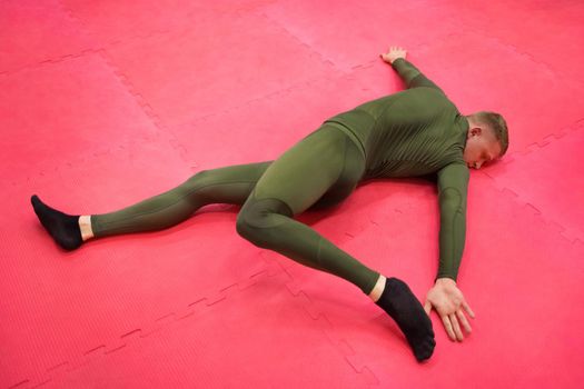 Young sports guy in a green thermal suit does exercises on the gym floor.