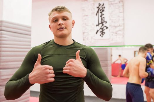 Young sports guy in a green thermal suit shows class against the background of a workout in the gym.