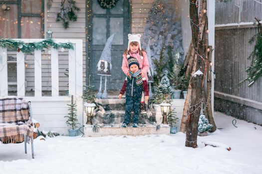 Happy little kids sitting on the porch of the Christmas decorated house, snowing outdoor. Happy New Year and Merry Christmas. Magic winter