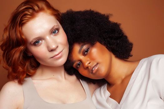 young pretty caucasian, afro woman posing cheerful together on brown background, lifestyle diverse nationality people concept close up