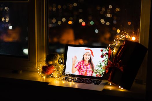 Laptop computer standing on table with gift and Santa hat in cozy room with little girl on the screen smiling, wishing Merry Christmas and Happy New Year.