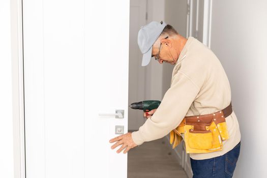 Attractive builder is installing lock in door. He is holding a screwdriver and kneeling