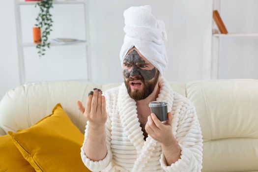 Bearded man having fun with a cosmetic mask on his face made from black clay. Men skin care and spa at home.