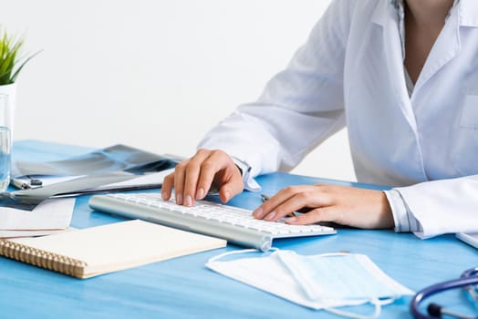 Close-up of female doctor hands typing at computer keyboard. Physician in medical uniform working at desk. Examination and diagnosis in hospital. Practitioner workplace with computer keyboard.