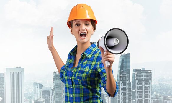 Expressive woman in safety helmet shouting into megaphone. Portrait of young emotional construction worker with wide open mouth on cityscape background. News announcement and advertisement.