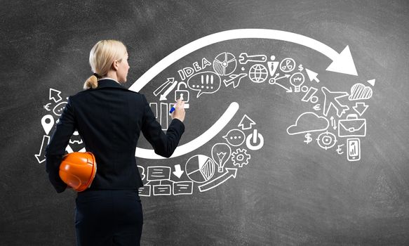 Businesswoman draws arrows consists of business doodles on blackboard. Back view woman with safety helmet and marker pen in hands. Operational strategy planning and project management concept.