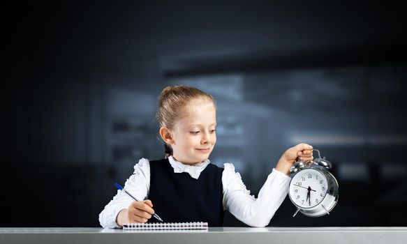 Smiling little girl holding alarm clock. Time to study and back to school concept. Happy schoolgirl sitting at desk with notebook and pen in hand. Time management. Research and education in school.