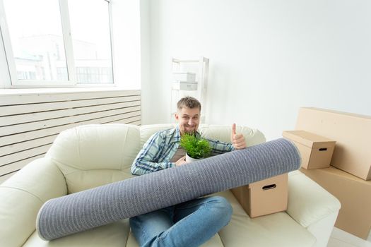 Positive young man sitting on the sofa with his things in the new living room after the move and shows a thumbs up. Concept of housing affordability for young people