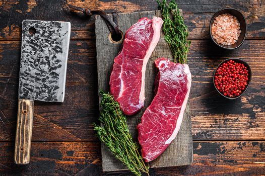 Raw sirloin beef meat steak on a wooden cutting board with herbs. Dark wooden background. Top view.