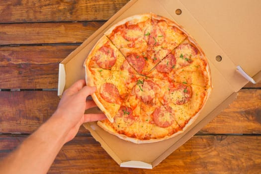 The guy's hand takes a pizza in cardboard box of against the background of a wooden table. Delicious fast food.