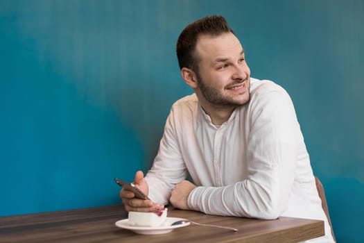 Portrait of a smiling businessman in a white shirt with a phone in his hands, having breakfast in a cafe on a blue background.