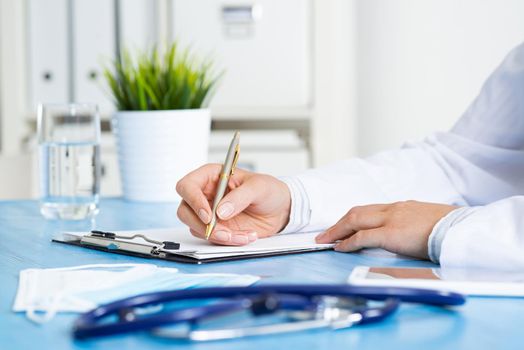Doctor filling patient history list at clipboard. Physician in medical uniform working at desk in office. Examination and diagnosis in clinic. Doctor workplace with green plant in pot and stethoscope.