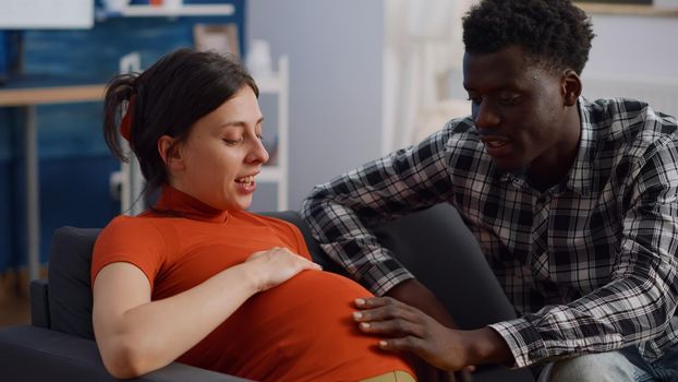 Close up of interracial couple with pregnancy at home holding hands on belly. Mixed race young partners expecting child and touching baby bump while sitting on living room couch