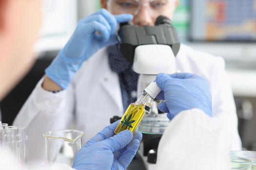 Close-up of persons hand hold jar with cannabis oil and going to add drop under microscope. Woman scientist explore sample, protective uniform. Lab concept