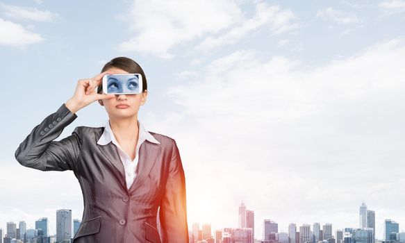 Portrait of pretty woman covering her eyes with smartphone. Businesswoman showing mobile phone with eyes on screen. Corporate businessperson on cityscape background. Mobile communication layout.