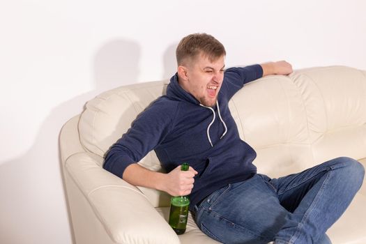 Depression and alcoholism concept - aggressive young man sitting on a sofa with bottle of alcohol.