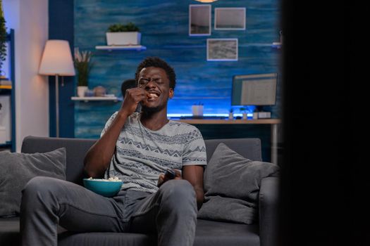 African american young man watching television laughing during comedy movie relaxing in living room. Black guy sitting on sofa while eating popcorn enjoying spending tine alone