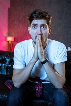 Young handsome man watching TV sitting on the sofa. Male fan of your favorite sports team. Emotional man in a white T-shirt